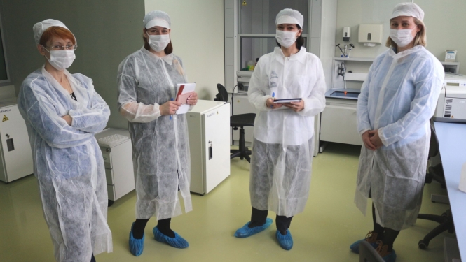Četras sievietes baltos halātos un ar maskām uz sejas stāv laboratorijas telpās